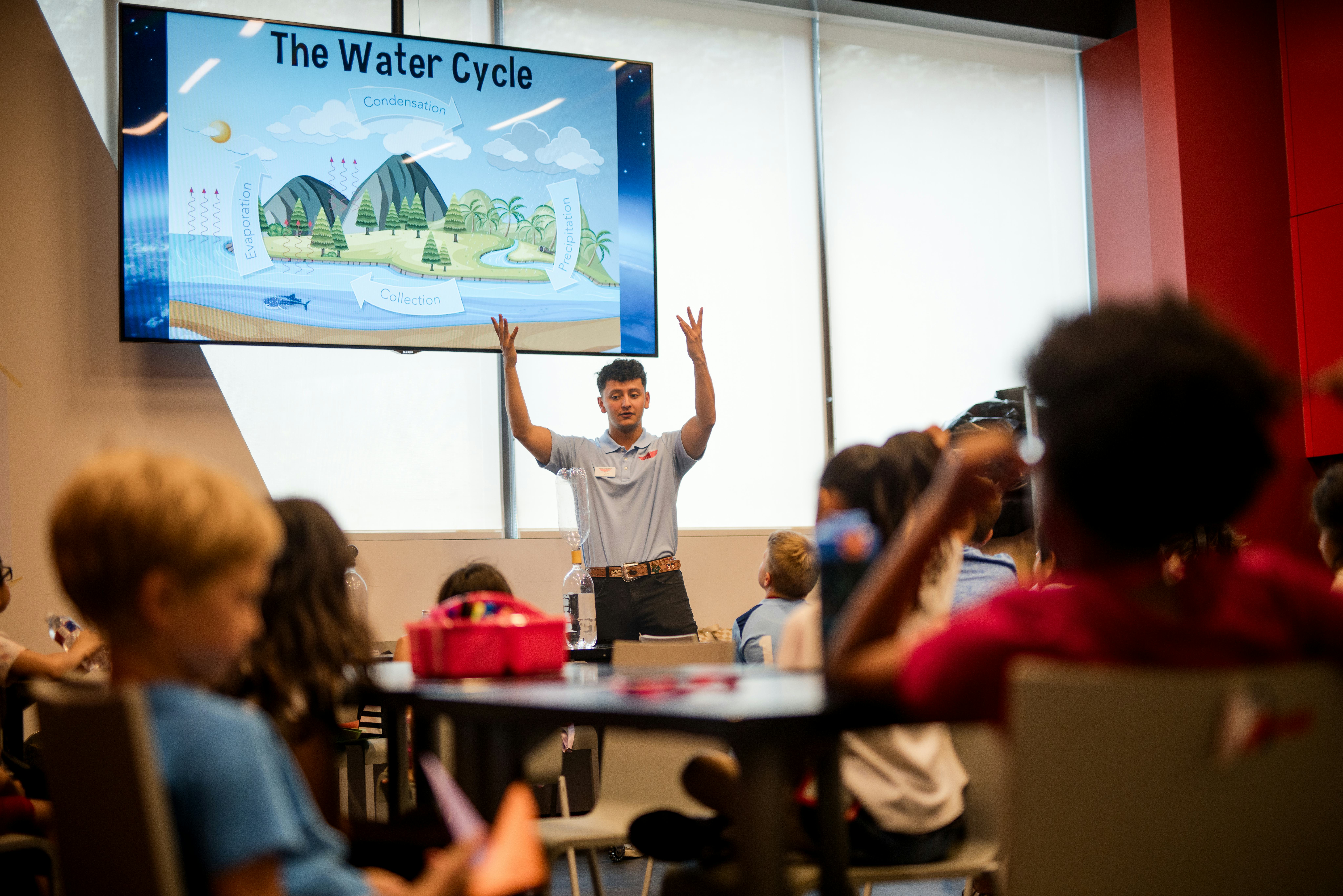 Adult standing with arms raised facing children seated in classroom setting; Adult standing in front of screen featuring 