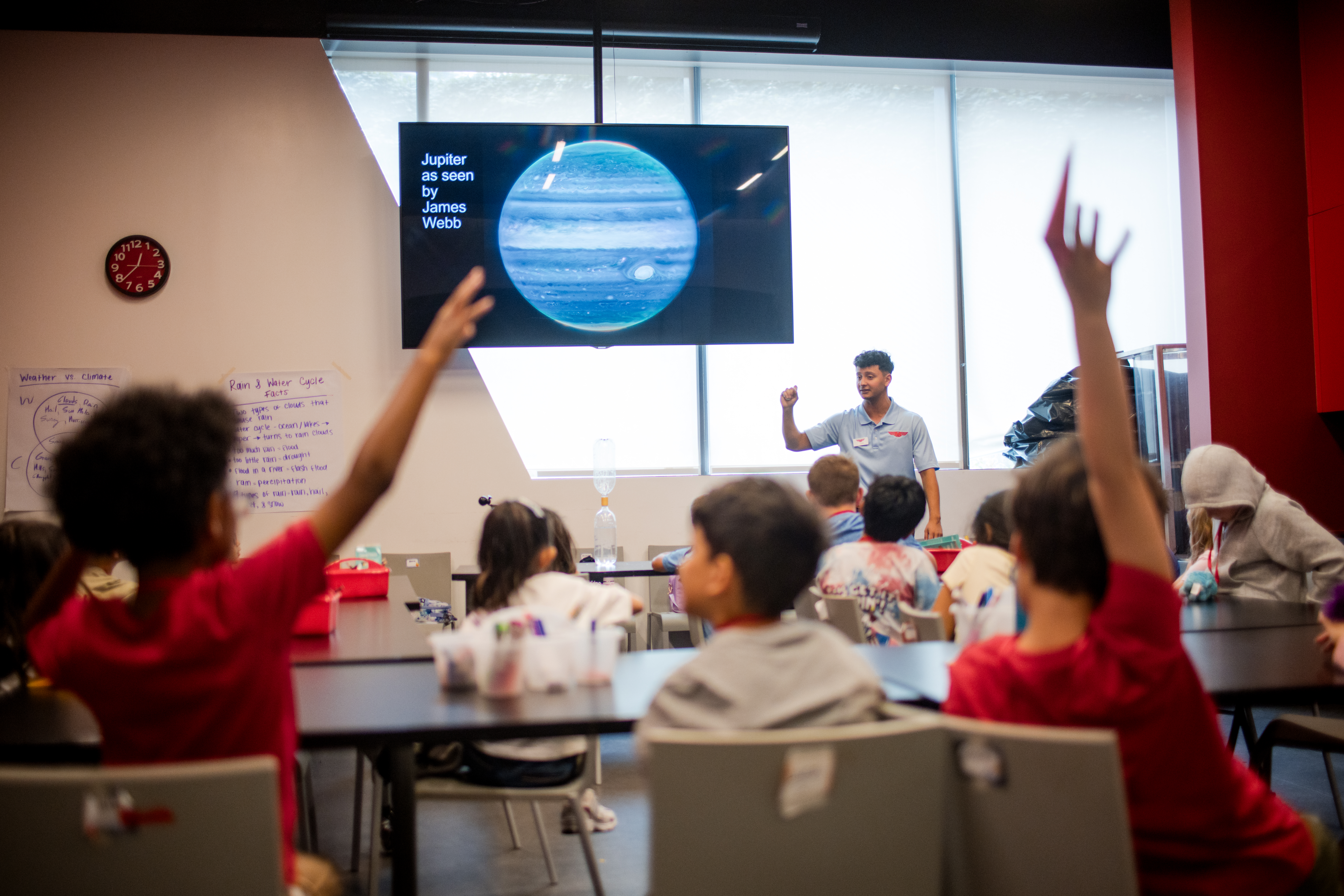 A group of seated children raising their hands in front of a man pointing toward tv screen
