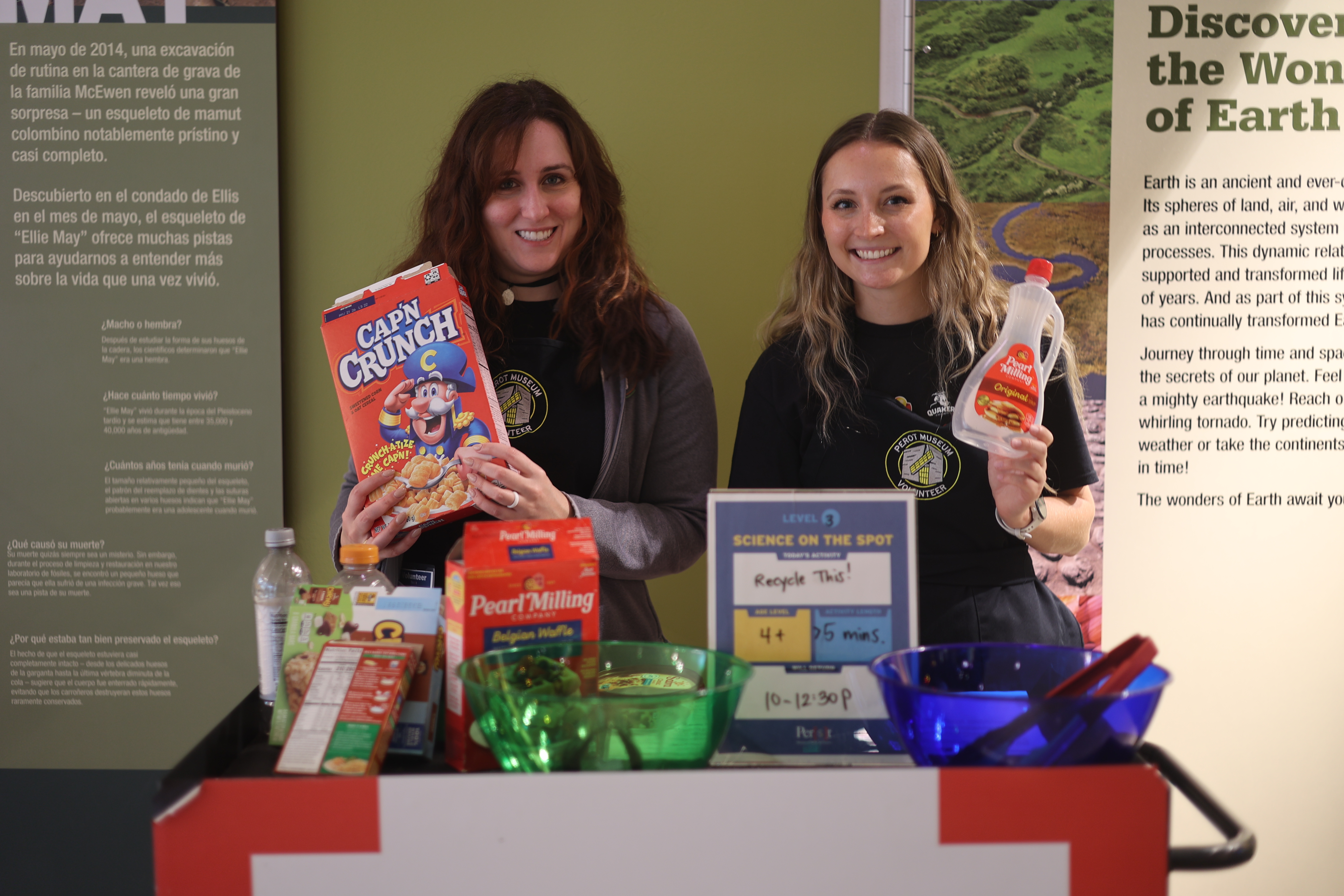 Two women smiling and holding a Cap'N Crunch box and bottle of maple syrup, strandig in front of a table with other snack-related items