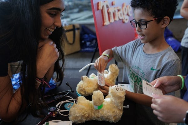 Little boy with glasses operating on teddy bear with adult looking and smiling