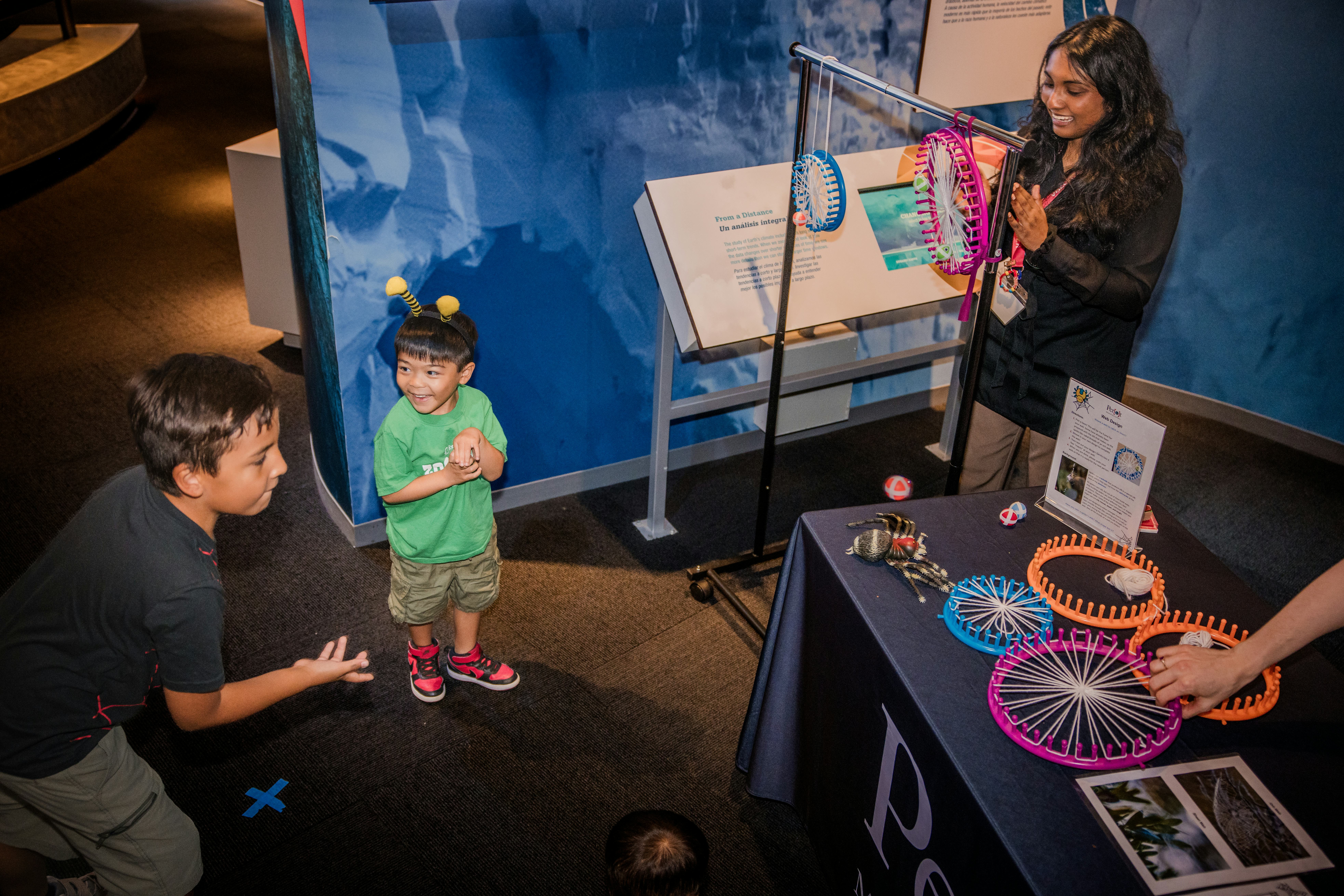 Older boy tossing ball onto activity table with younger boy smiling at him, both in front of adult woman smiling and attempting to clap
