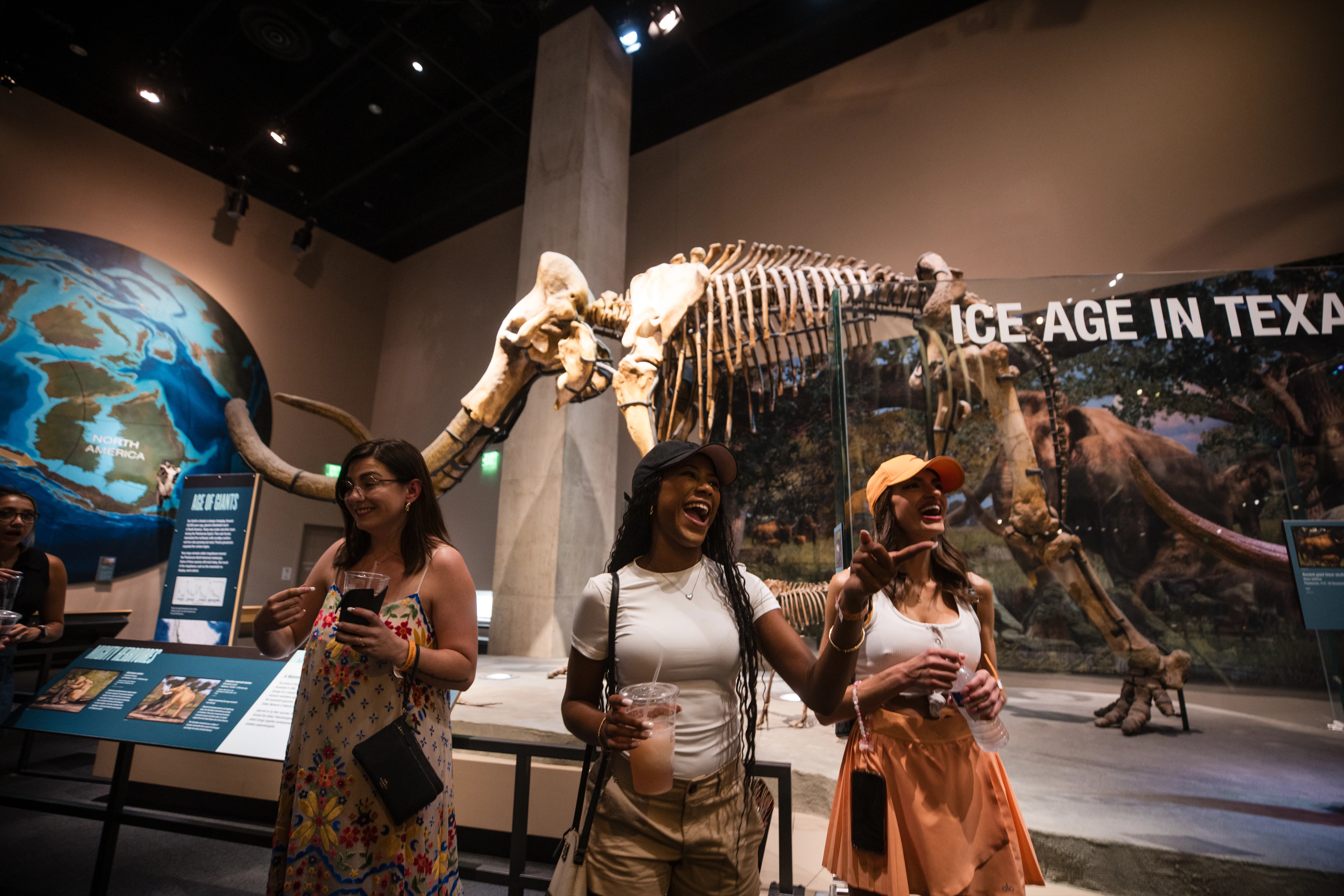 Three women smiling and holding drinks in front on dinosaur fossil