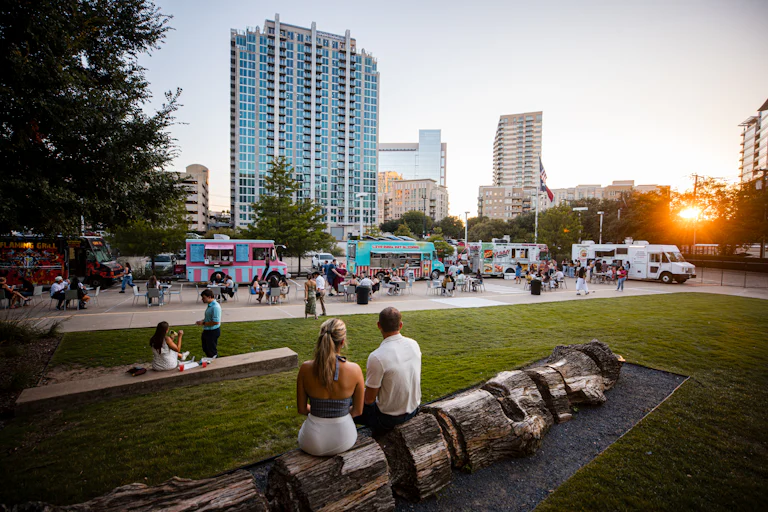 Man and woman sitting on log looking toward line of food trucks and groups of people
