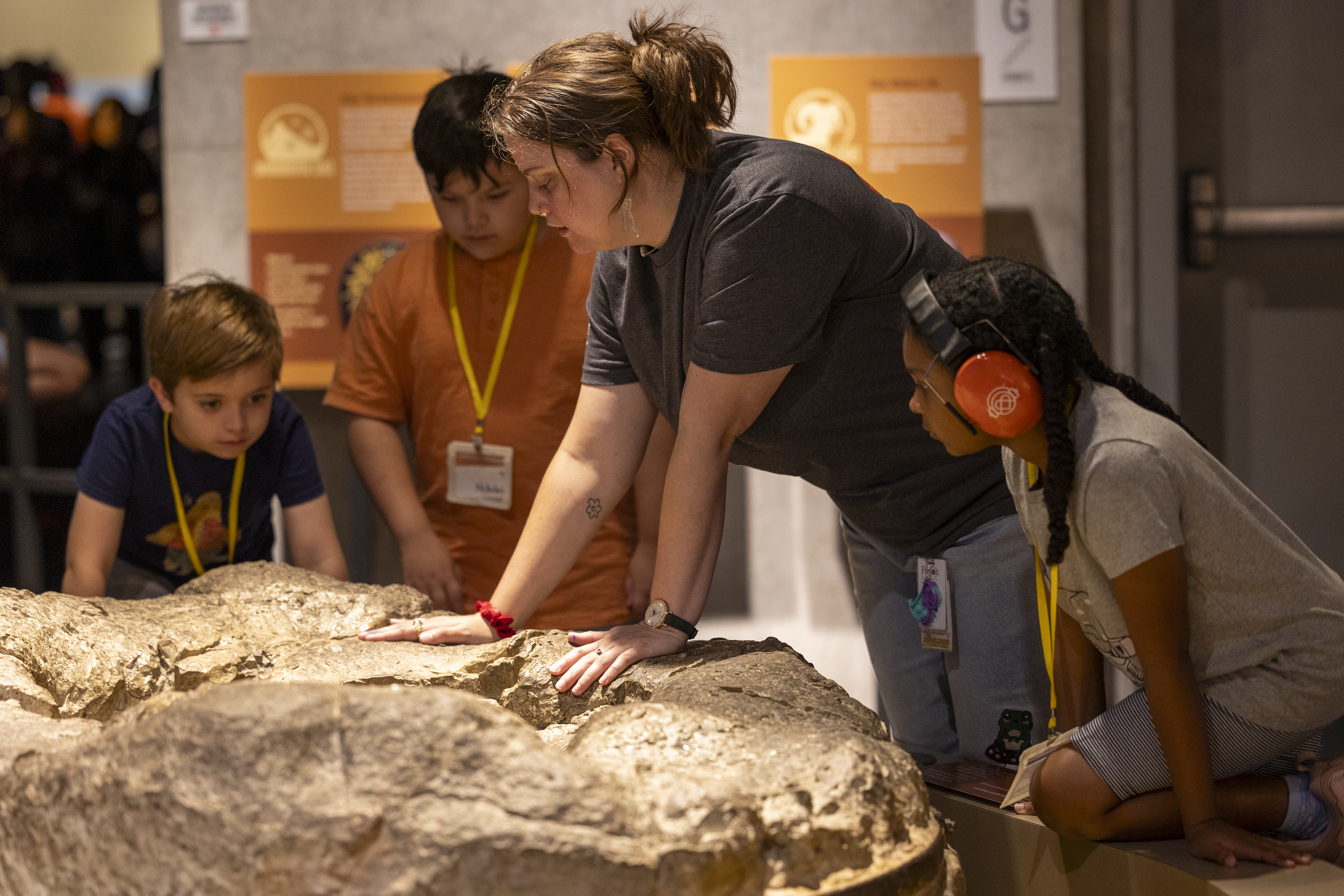 Young girl wearing sensory headphones with two other children and adult woman staring down at large rock exhibit